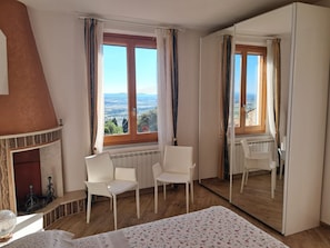A bedroom with a fireplace, two chairs, a bed, a large mirror, and a view of the countryside.