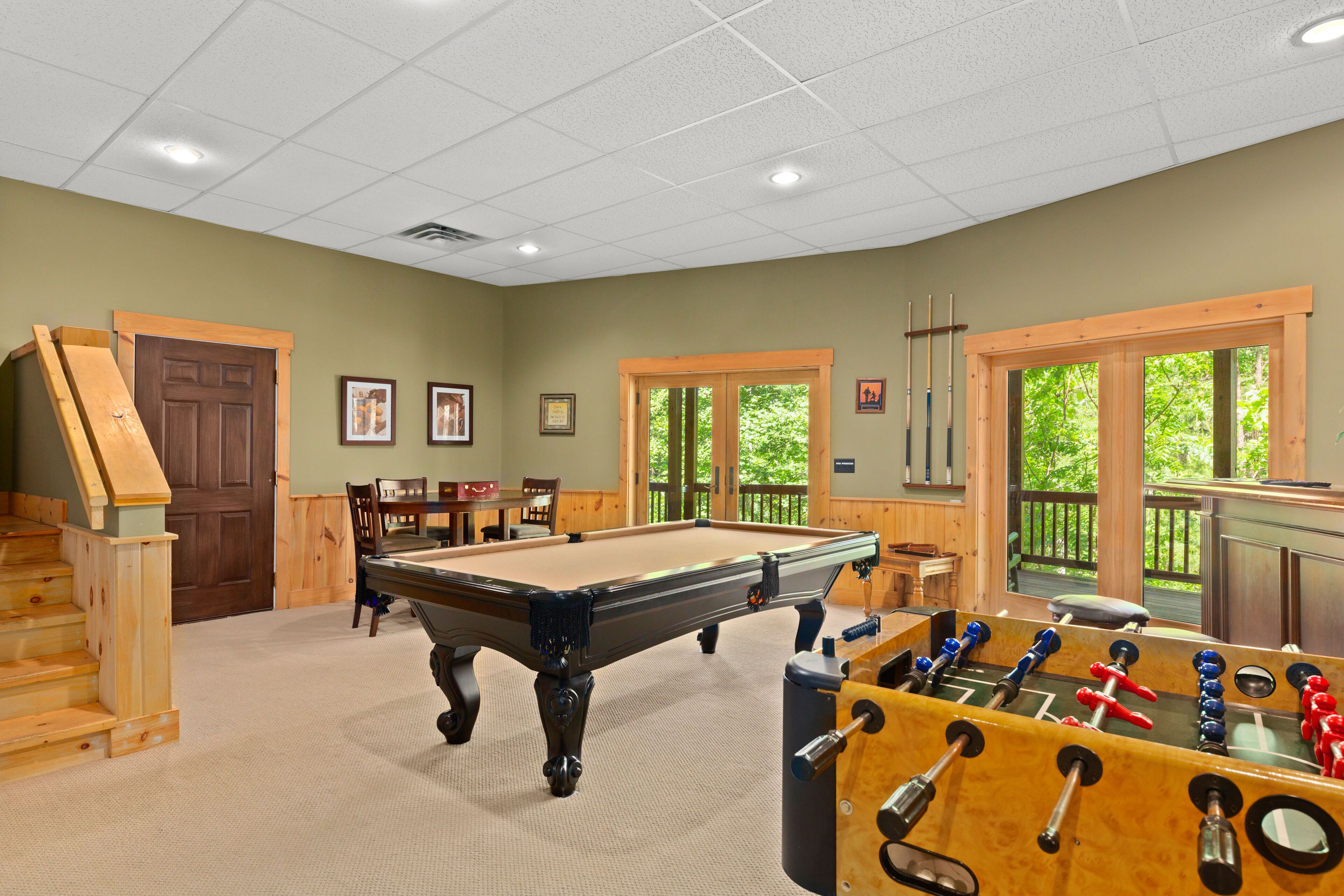 A pool table, foosball table, and a staircase in a room with green walls and windows.