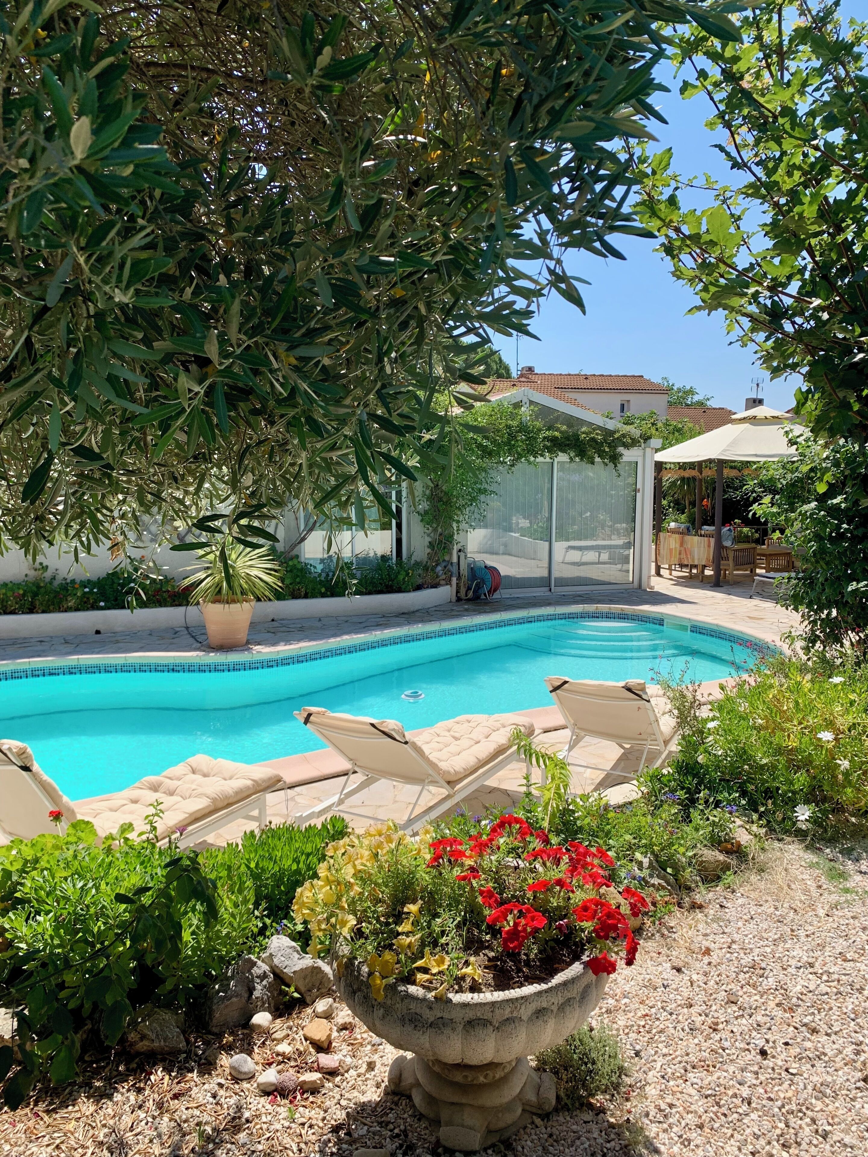Une piscine avec des chaises longues, un patio couvert et un jardin agrémenté d’un grand pot de fleurs.