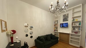 A living room with a dark green sofa, a wooden floor, a chandelier, a TV on the wall, and a bookshelf.