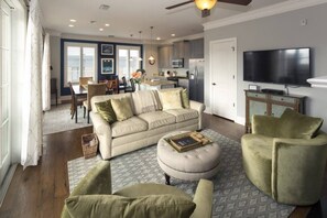 Una sala de estar amplia con sofá, reposapiés y sillón, un comedor con sillas, una cocina equipada y un televisor montado en la pared.