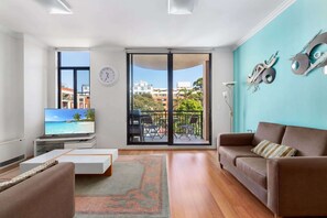 A living room with a TV, sofa, coffee table, and a view of buildings through a sliding door.