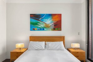 Une chambre à coucher avec un lit en bois, du linge de lit blanc, deux lampes de chevet et un tableau abstrait aux couleurs vives accroché au mur.