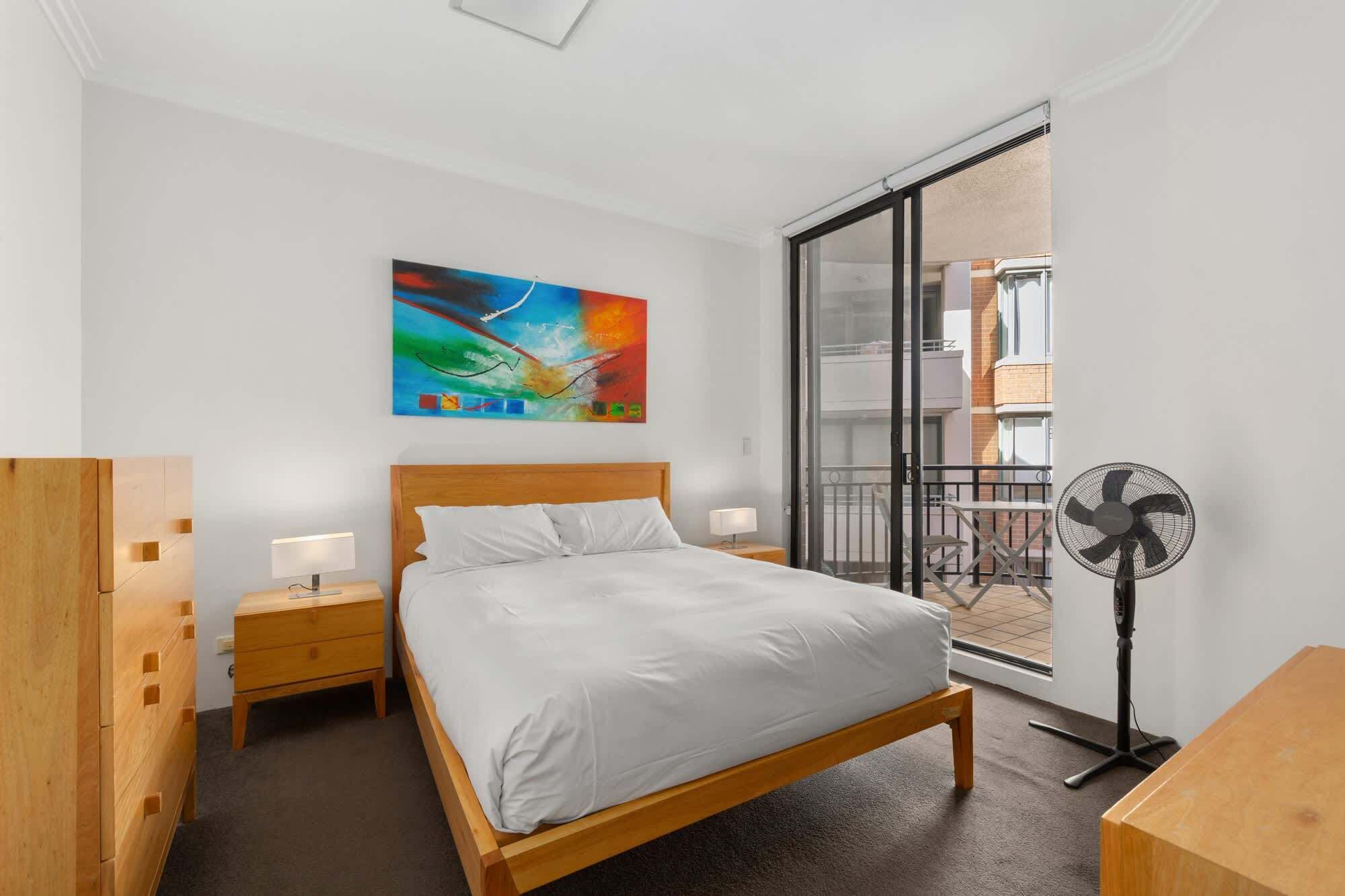 Een slaapkamer met een bed, nachtkastje, kapstok, ventilator en een groot raam met uitzicht.