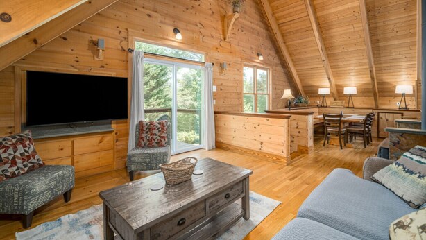 Un acogedor interior de cabaña de madera con una televisión grande, asientos cómodos, una zona de comedor y una cocina.