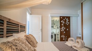 Un dormitorio con una cama, cabecera de madera, una puerta blanca, y un baño visible a través de una puerta de cristal. Se aprecia un perchero con toallas.