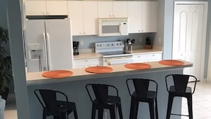 Eine moderne Küche mit weißen Schränken, einer Mikrowelle und einem Herd. Vier schwarze Barhocker und ein weißer Kühlschrank sind vorhanden.