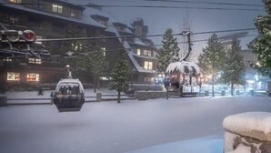 Een besneeuwde straat met een bewegende kabelbaan, gebouwen met traditionele architectuur en een stenen muur.