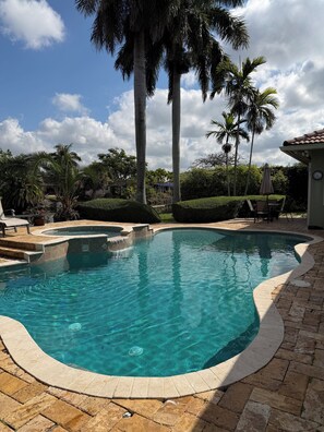 A clear swimming pool with a tiled edge, surrounded by palm trees and a brick patio.