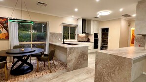A modern kitchen with a marble countertop, a dining area with a round table, and a view of greenery outside. - Luxury 5BR Lakefront Homestead — 55 Acres, Sandy Beach, Firepit (Oberon)