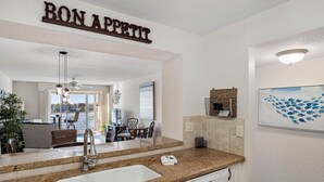 A kitchen with a granite countertop, white cabinets, and a sink. There is a ceiling fan, a chandelier, and a dining area visible in the background.