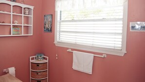 Un baño rosa con un estante blanco, un cuadro, una ventana con persianas y un perchero.