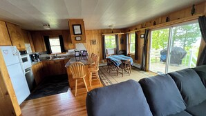 Un intérieur de chalet chaleureux comprenant une cuisine, un coin repas et une grande fenêtre donnant sur un paysage verdoyant.