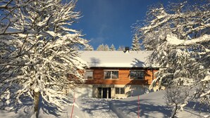 A wooden cabin with a snow-covered roof, surrounded by snow-laden trees, and a clear blue sky.