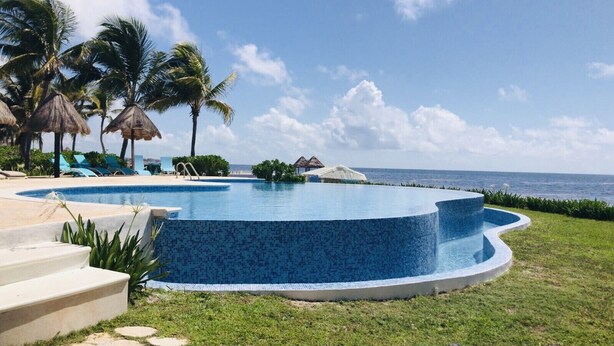 Ein kreisförmiger Infinity-Pool mit blauen Mosaikfliesen, umgeben von einer Rasenfläche, Palmen und Liegestühlen.