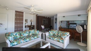 A living room with two sofas, a coffee table, a ceiling fan, a kitchen area with a microwave, and a refrigerator.