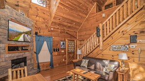 Interior acogedor de una cabaña de troncos con chimenea de piedra, muebles de madera, un televisor montado en la pared y una escalera que conduce a un nivel superior.