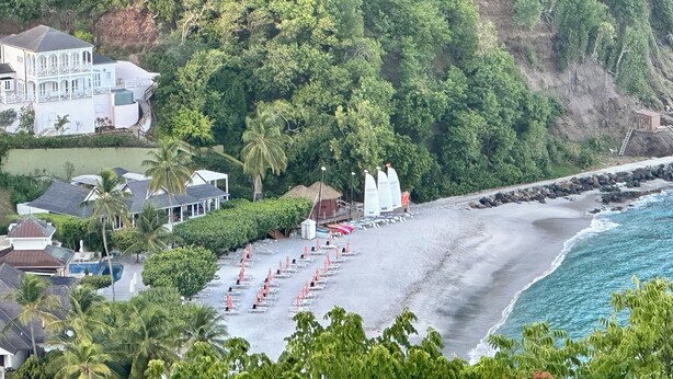 Un hotel frente al mar con playa, un edificio grande con varios balcones y un acantilado rocoso.