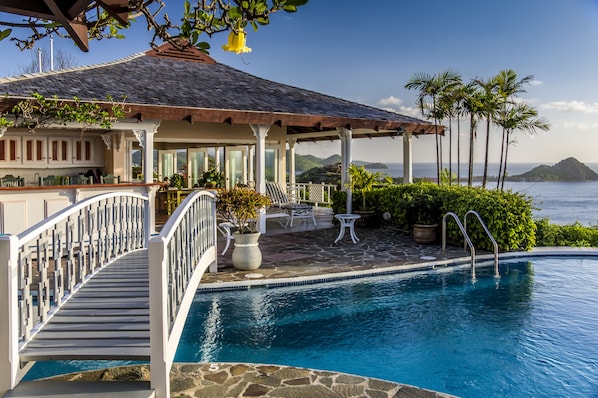 A pool area with a covered patio, lounge chairs, and a view of the ocean.