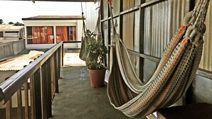 A balcony with a hammock, a potted plant, and a view of a building with red curtains. - Hostel Trotamundos (Alajuela)