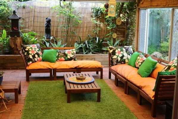A cozy outdoor seating area with two wooden sofas and a small table, surrounded by lush greenery and a bamboo fence. - A Great Family Place in Henley Beach - Pets  ✅ opposite park stylish & comfy (Henley Beach)