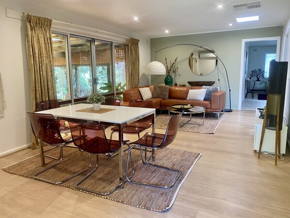 A modern living room with a dining area, featuring a glass table, leather sofa, and a large mirror.