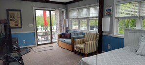 A room with a bed, a sofa, a striped armchair, a TV, a table, and a window with blinds.
