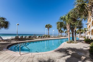 Une piscine à bord incurvé, entourée de chaises longues et de palmiers, avec une plage et l’océan en arrière-plan.