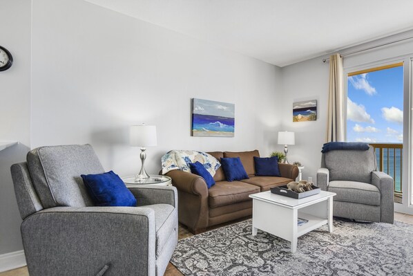 A living room with a grey sofa, a grey armchair, a white coffee table, and a patterned rug. There are two lamps, a painting of a beach, and a window with a view of the ocean. - 1PM Early Check In! Beachfront! Walk to Pier Park!  Beach Chair Service included (Panama City Beach)