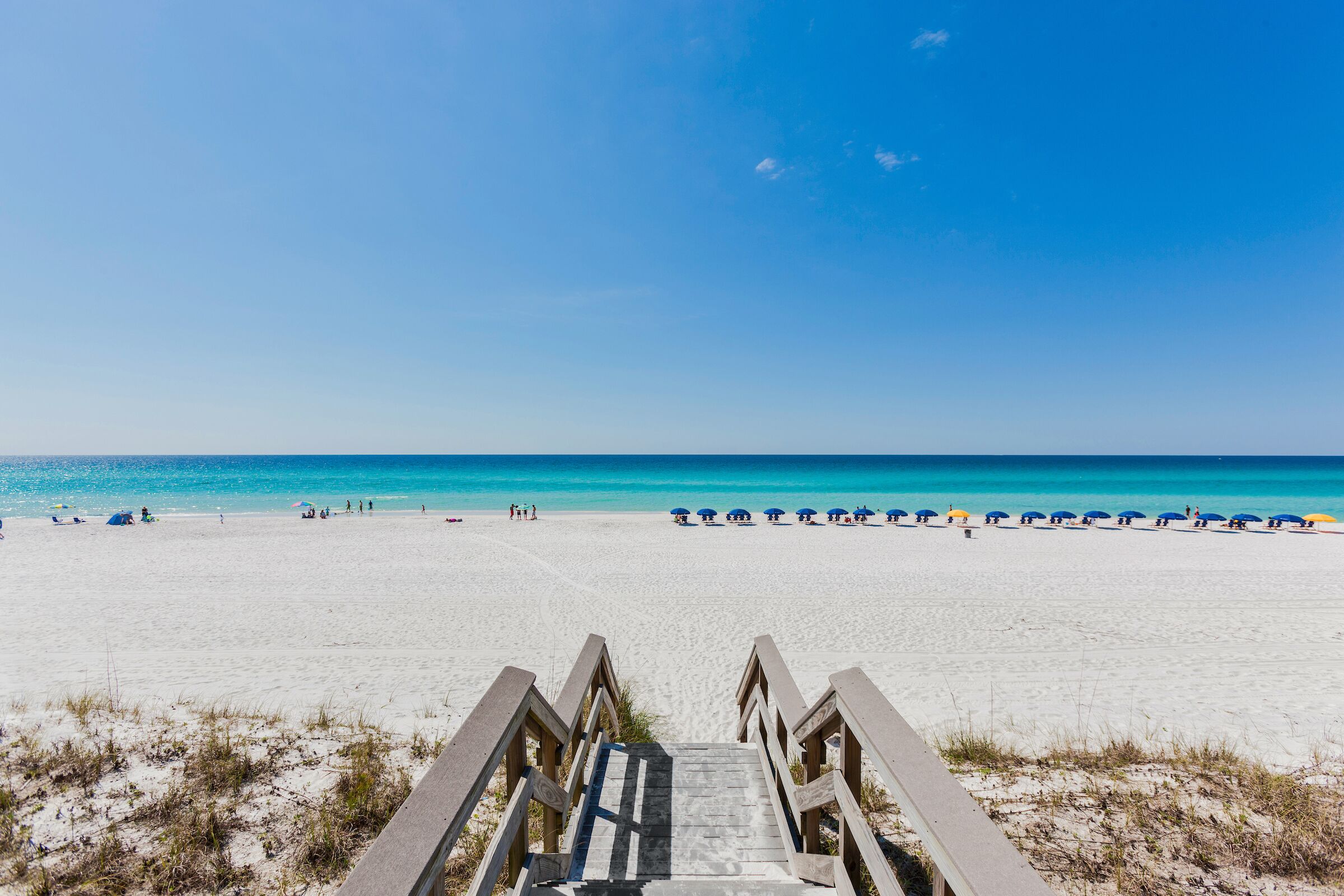 Una spiaggia con una scala di legno che conduce alla sabbia, un cielo azzurro limpido e un mare calmo.