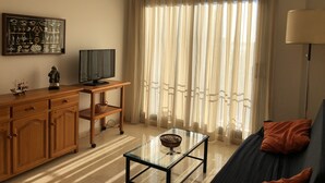 A living room with a sofa, a glass coffee table, a TV on a wooden stand, and a large window with sheer curtains. - COZY APARTMENT 50 M FROM THE BEACH with WIFI (L'Ampolla)