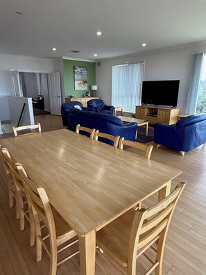 A spacious living area with a wooden dining table and blue sofas, a television, and a kitchen in the background.