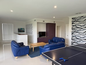 A modern living room with a ping pong table, blue sofas, a wooden coffee table, and a TV.