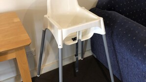 A white high chair with four legs, positioned next to a wooden table and a dark blue couch with a patterned cover.