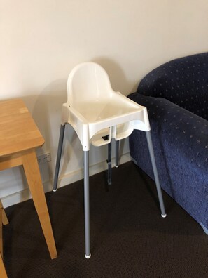 A white high chair with four legs, positioned next to a wooden table and a dark blue couch with a patterned cover.