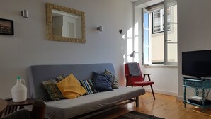 A living room with a sofa, a red chair, a television, and a window with a view of buildings. - Historical apartment in Alfama (Lisboa)