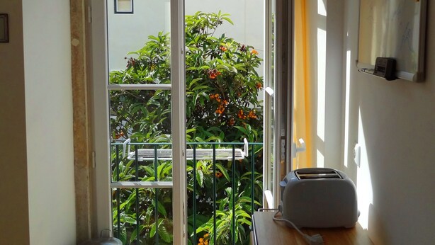 A room with a sliding glass door leading to a balcony with plants and a view of buildings. There is a trash can, a table with a toaster, and a whiteboard on the wall. - Historical apartment in Alfama (Lisboa)