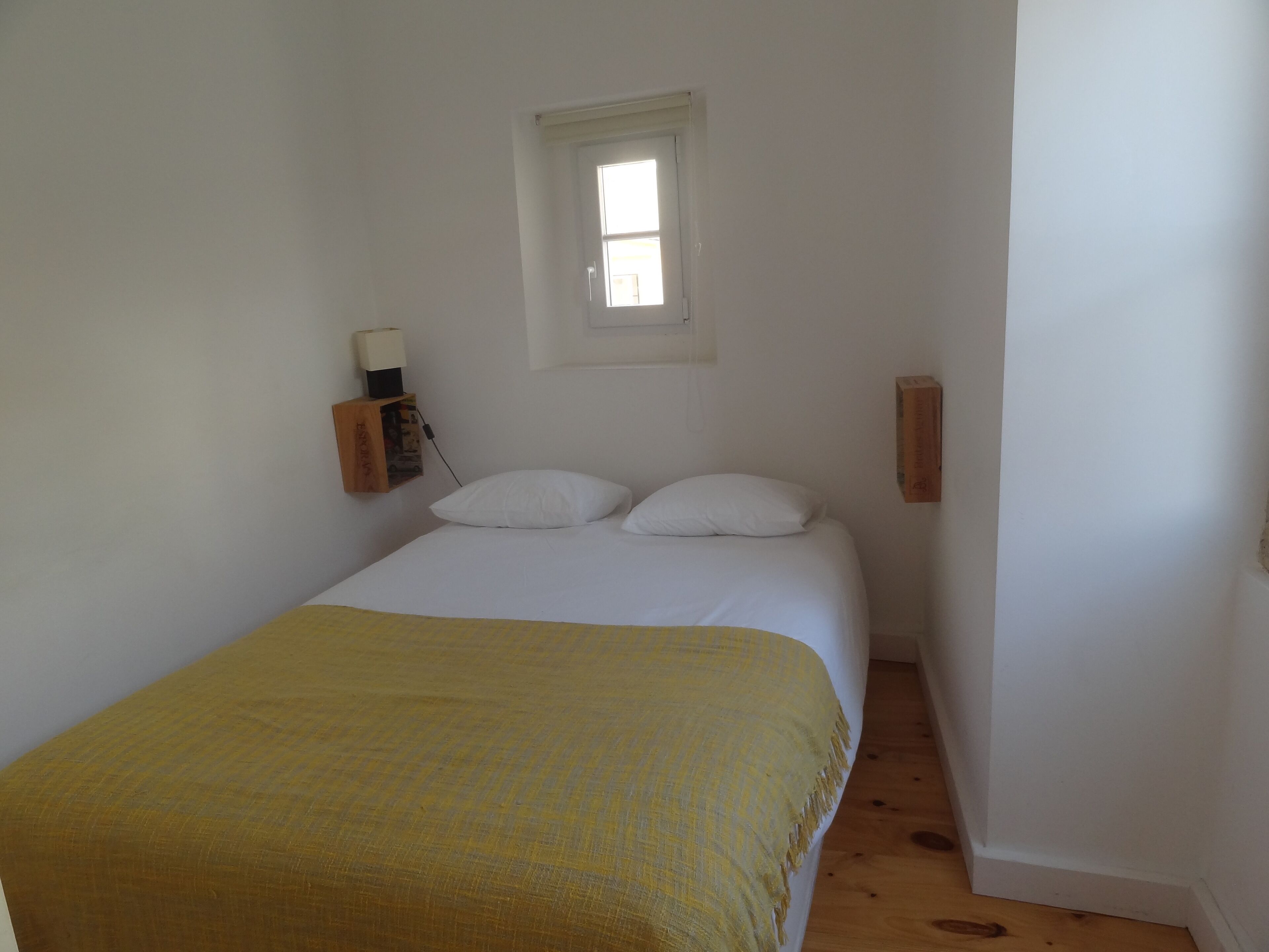 A small, simple bedroom with a bed, two pillows, a bedside table with a lamp, and a window.