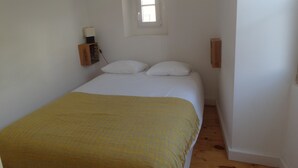 A small, simple bedroom with a bed, two pillows, a bedside table with a lamp, and a window. - Historical apartment in Alfama (Lisboa)