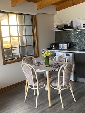 A dining area with a table and four chairs, a window with a view, a kitchen area with appliances, and a green vase with flowers on the table.