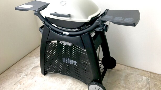 Una griglia portatile con piano superiore bianco e base nera, caratterizzata da un pannello laterale in metallo forato e una manopola di controllo.