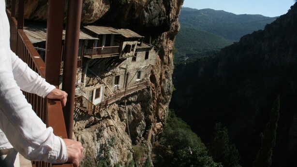 Un bâtiment rustique en pierre, perché sur une falaise, avec un balcon en bois donnant sur une vallée.