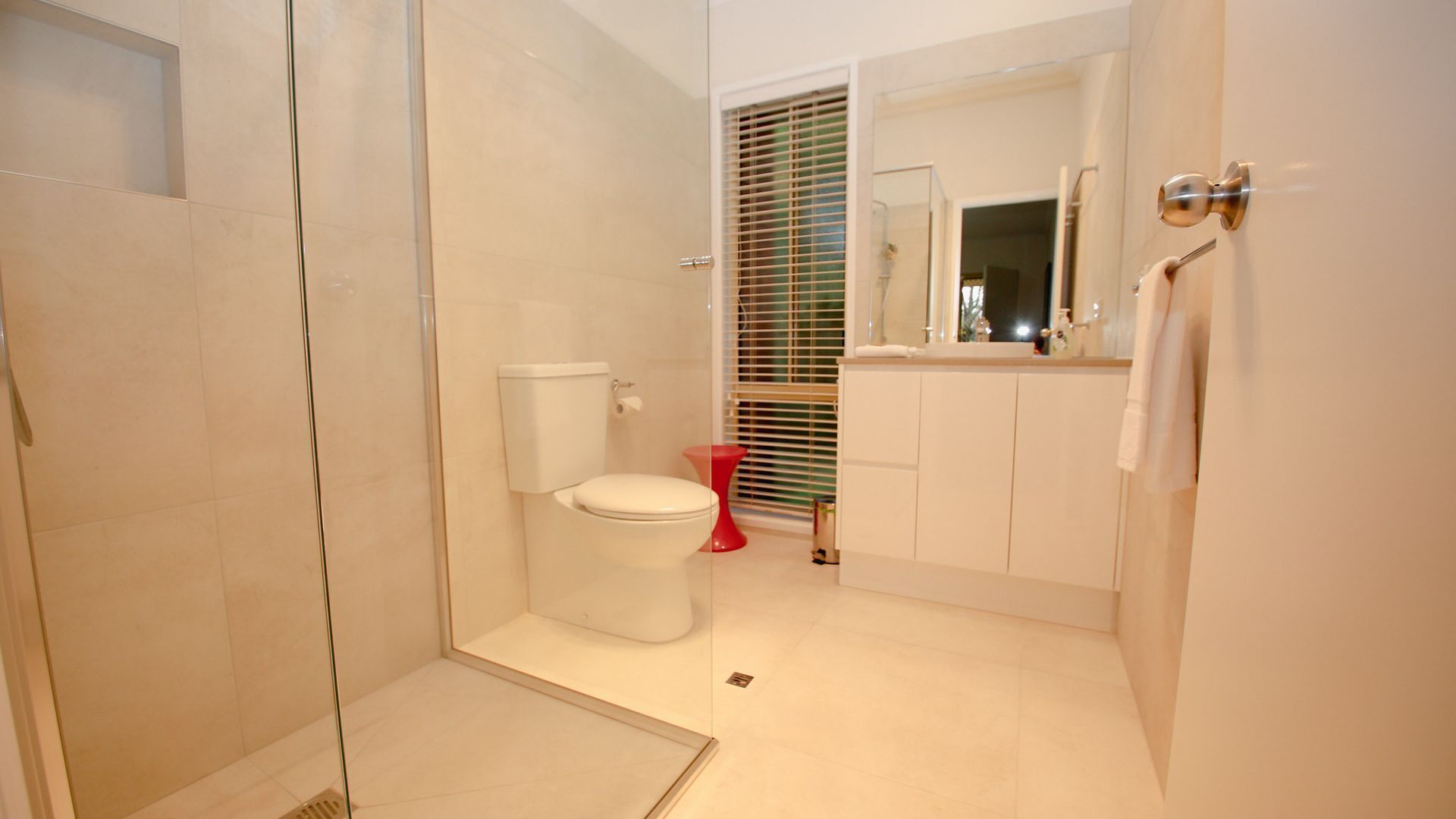 Un bany modern amb un espai de banyera de vidre, un vàter, un lavabo amb mirall i una finestra amb persianes.