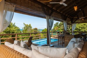 Un área de estar al aire libre cubierta con piscina, mobiliario de madera y vistas al océano.
