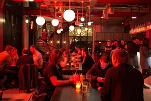 A crowded bar with people sitting at tables, a staircase in the background, and hanging spherical light fixtures.