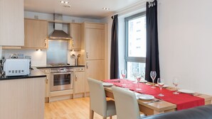 A modern kitchen with wooden cabinets, a built-in oven, and a dining area with a red tablecloth. - Providing Stylish City Living within the vibrant shore area (Edinburgh)