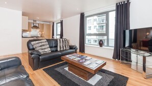 A modern living room with a leather sofa, a coffee table with magazines, a glass TV stand, and a large window with curtains. - Providing Stylish City Living within the vibrant shore area (Edinburgh)