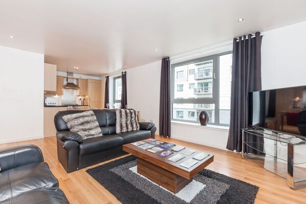 A modern living room with a leather sofa, a coffee table with magazines, a glass TV stand, and a large window with curtains.