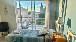 Ein Schlafzimmer mit zwei Betten, einem Sessel, einem Nachttisch mit Lampe und Blick auf Gebäude durch ein großes Fenster.