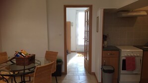 A dining area with a glass table and wicker chairs, a kitchen with a stove and a trash bin, and a door leading to another room. - 
Gite with pool & fab gardens 5 mins from quaint village with restaurants & shop (Trémolat)